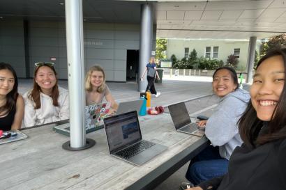 In this photo, there are five girls sitting at a table and posing for a selfie. Three girls sit on one side of the table and two girls sit on the other side. Four computers are open on the table.